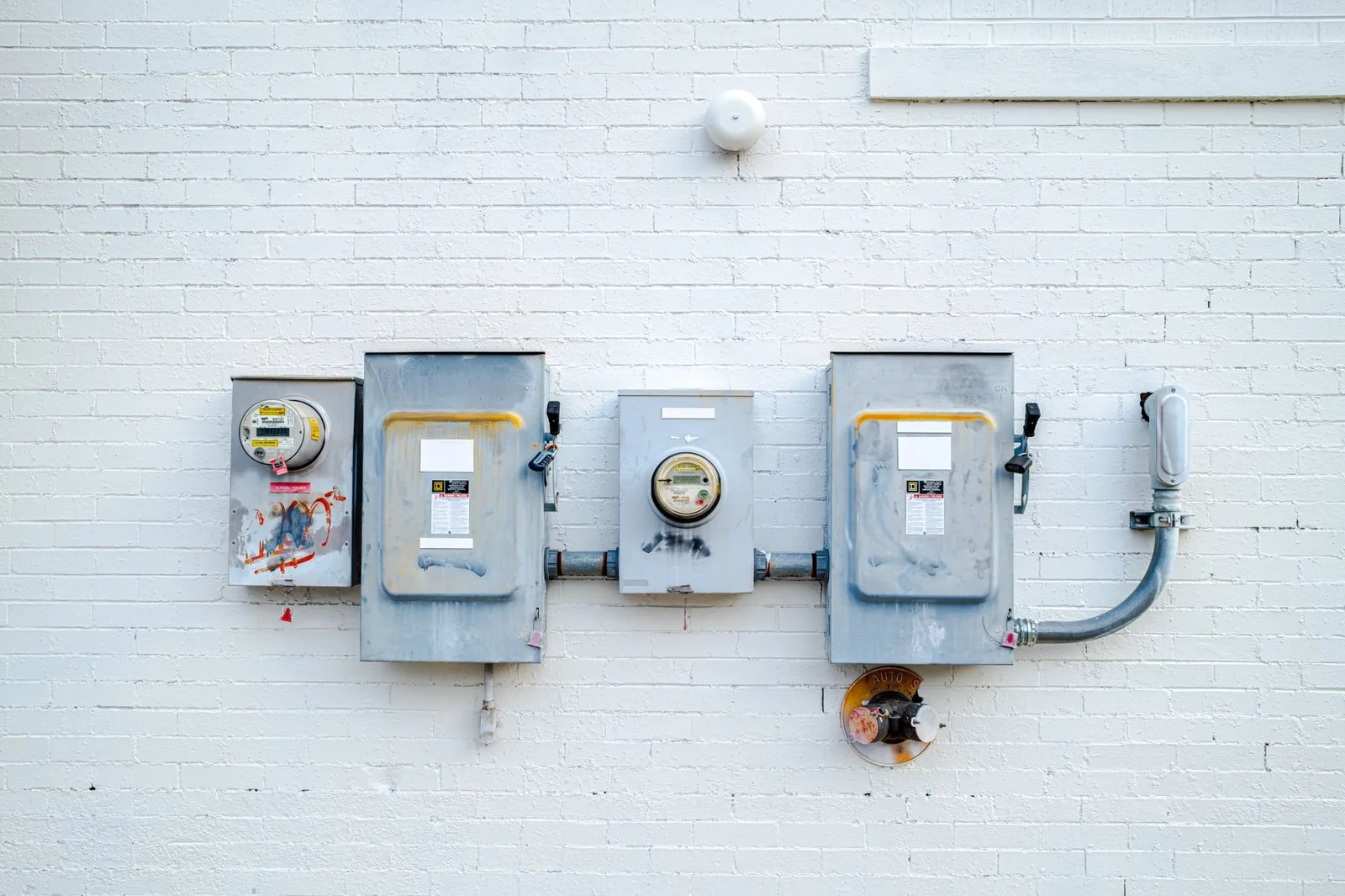 Two utility meters and two grey lockable safety switch disconnects mounted in a row on a white painted brick wall, linked by rigid metal conduit.
