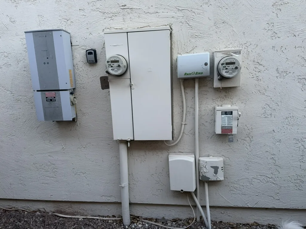 A residential exterior wall with two round utility meters in their sockets, a combination panel enclosure, a lockable disconnect, and white conduit running down to grade.