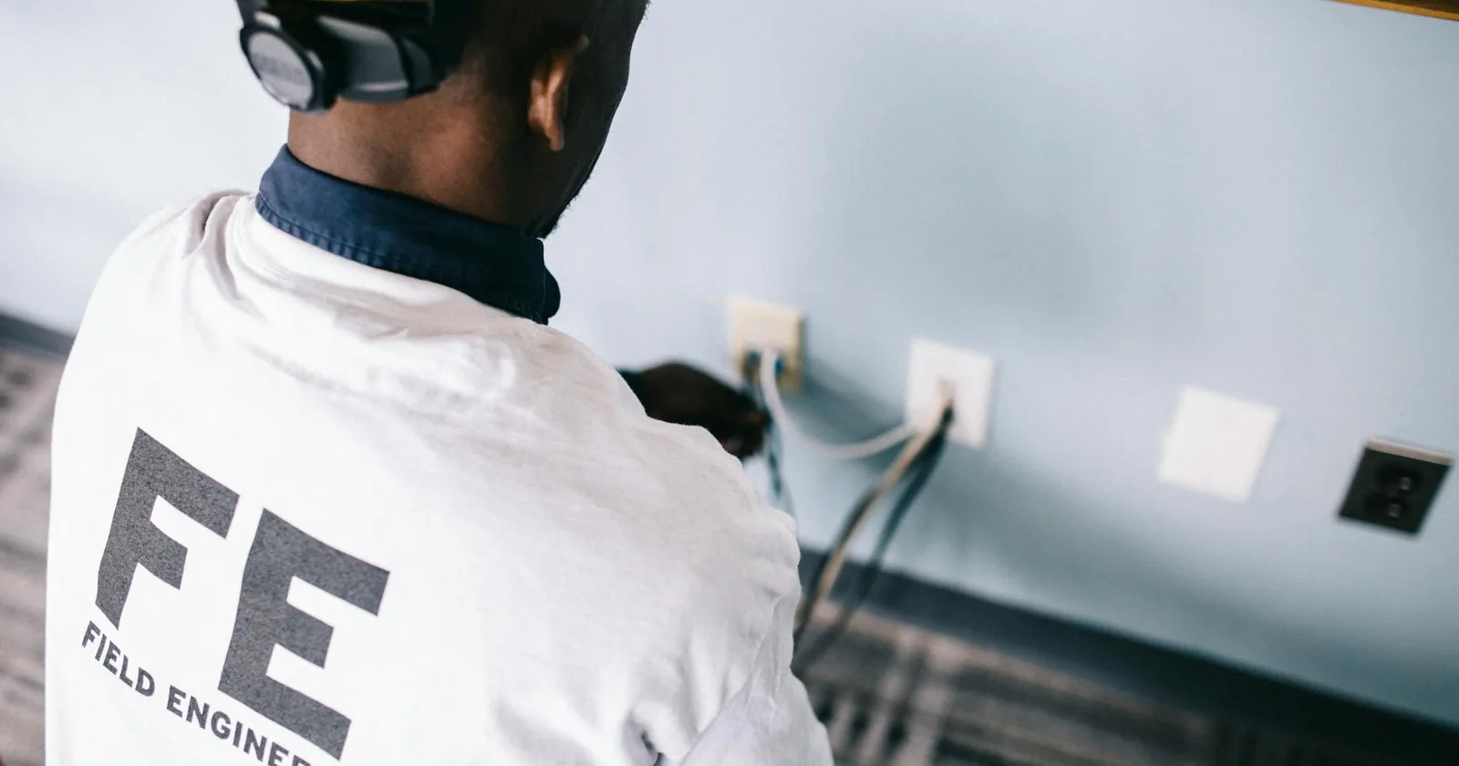 Field engineer in a white shirt and hardhat checking voltage at a wall outlet.
