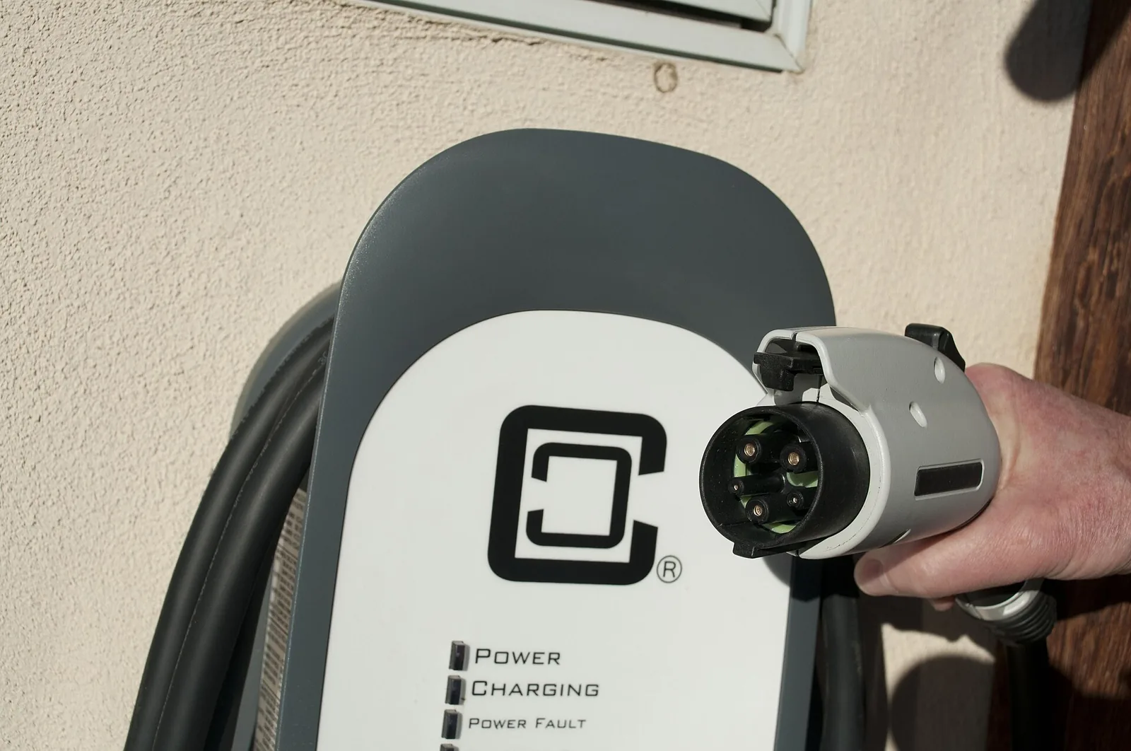 A hand holding a J1772 charging plug beside a wall mounted charger