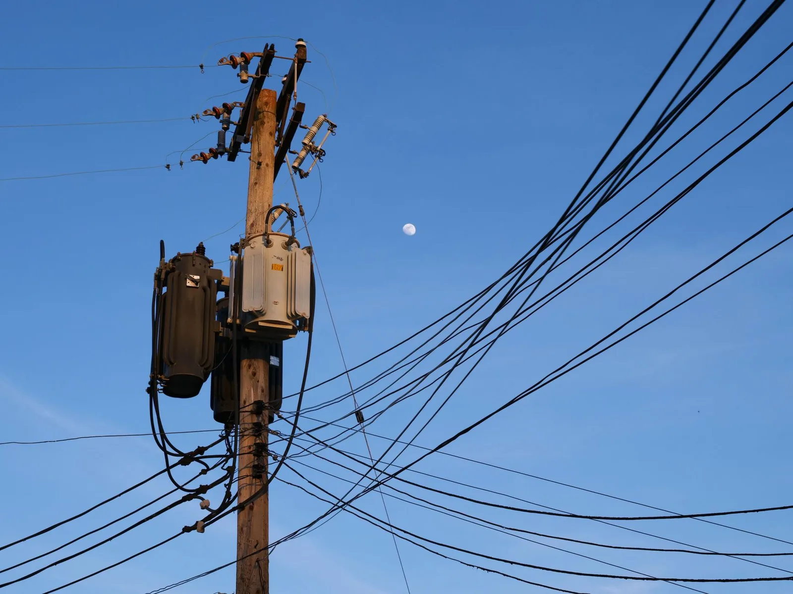 A wooden utility pole carrying pole-mounted transformers, cutout fuses, and a dense fan of overhead service lines against a blue sky.